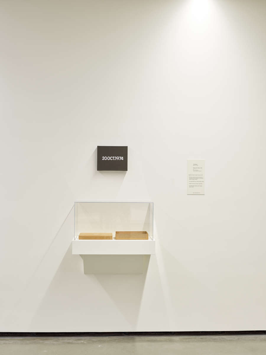  An installation photo depicting part of a white gallery room. A small black panel with illegible text is off-center on the wall, below, an enclosed shelf holds two rectangular objects.