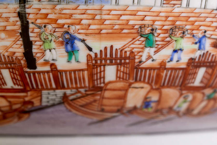 A close up of a painted ceramic depicting docked wooden boats in the foreground and workers carrying goods on an orange-tiled dock.