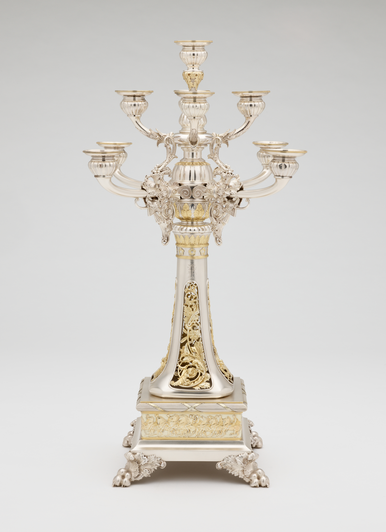 Front view of a silver nine branched candle holder, with detailed gold and ivory ornamentation of flowers and men on horseback.