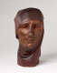 Front view of a vibrant, carved wooden sculpture depicting a serene face wearing a cloth draped around its head. The bust features smooth contours and a crack alongside its nose.