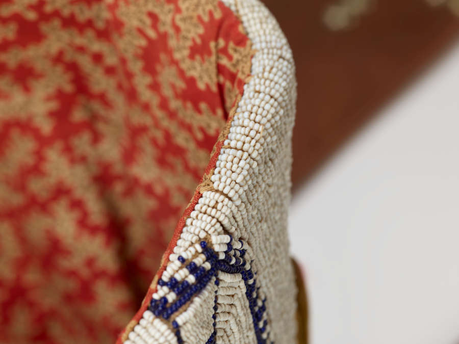 Detail of an object. Portion at left is a dense cream and red floral print. At right is a section covered in tiny blue and white beads.
