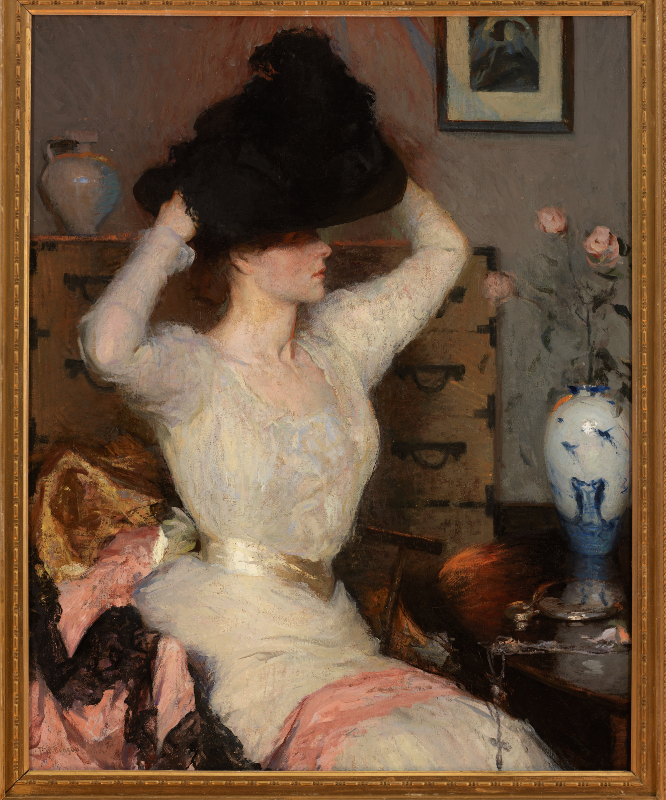 A woman in a white gown places a large black hat on her head inside a room decorated with pink flowers and a cabinet. 