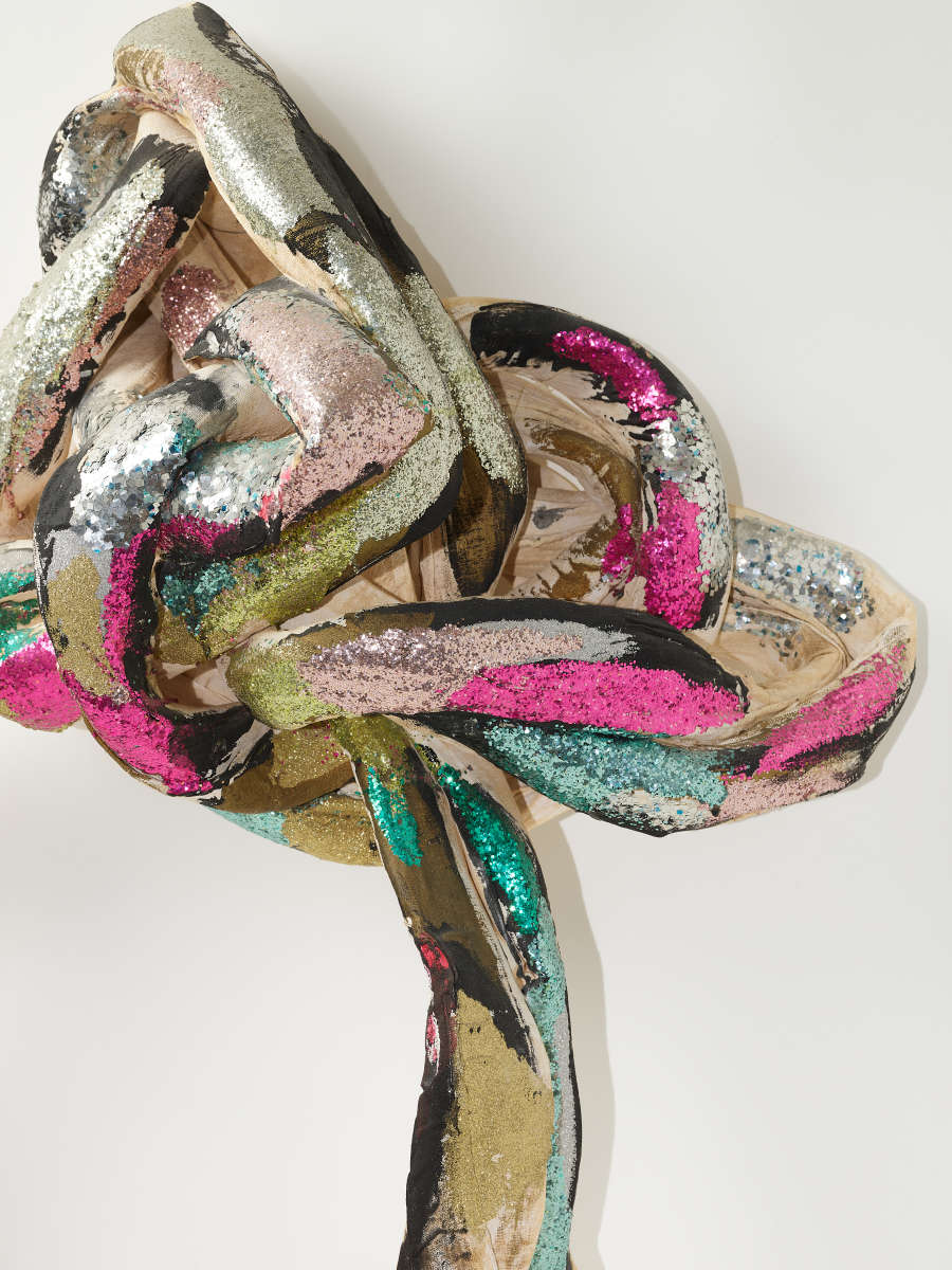 A knotted bundle of textile strips with metallic silver, pink, green, and black cloth, overlapping in a shimmering tangle.