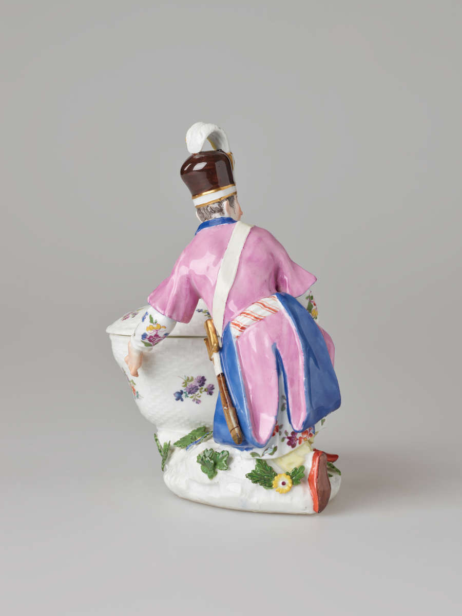 A sculptural figure kneeling next to a basket. Figure wears bright, elaborate clothing with a hat with a feather. There are floral decorations on the figure’s clothing and basket.
