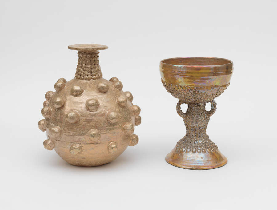 Two metallic vessels: one rounded with raised spherical studs and a narrow neck, the other a goblet with a shimmering bowl on a textured stem and small, looped handles.