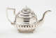 A silver teapot with a decorative angular handle, a rounded square body and spout with sculptural decorations.