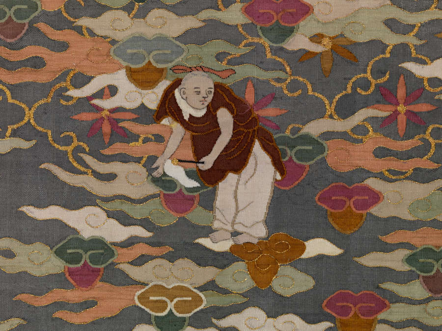Section of illustrations of the blue robe’s front featuring intricate gold and red floral patterns overlaid with wispy pastel clouds and a bent robed monk holding a fly whisk.