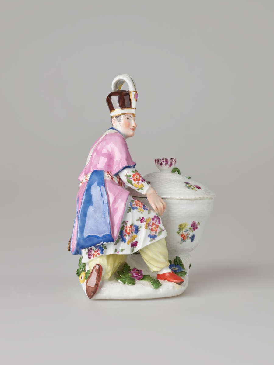 A sculptural figure kneeling next to a basket. Figure wears bright, elaborate clothing with a hat with a feather. There are floral decorations on the figure’s clothing and basket.
