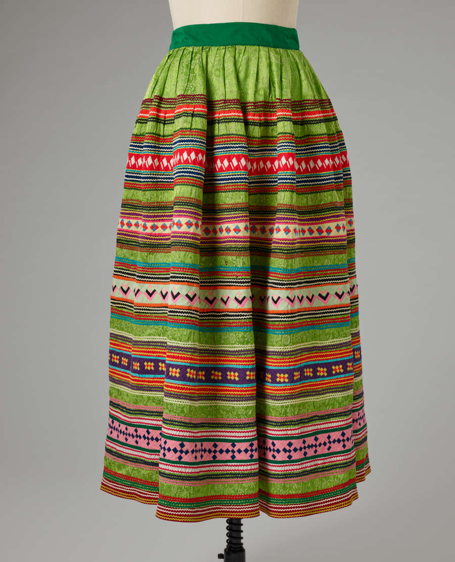 Green skirt on a mannequin, which is patterned with horizontal, warm and cool stripes, often containing patterns within them. The waistband is dark green. 