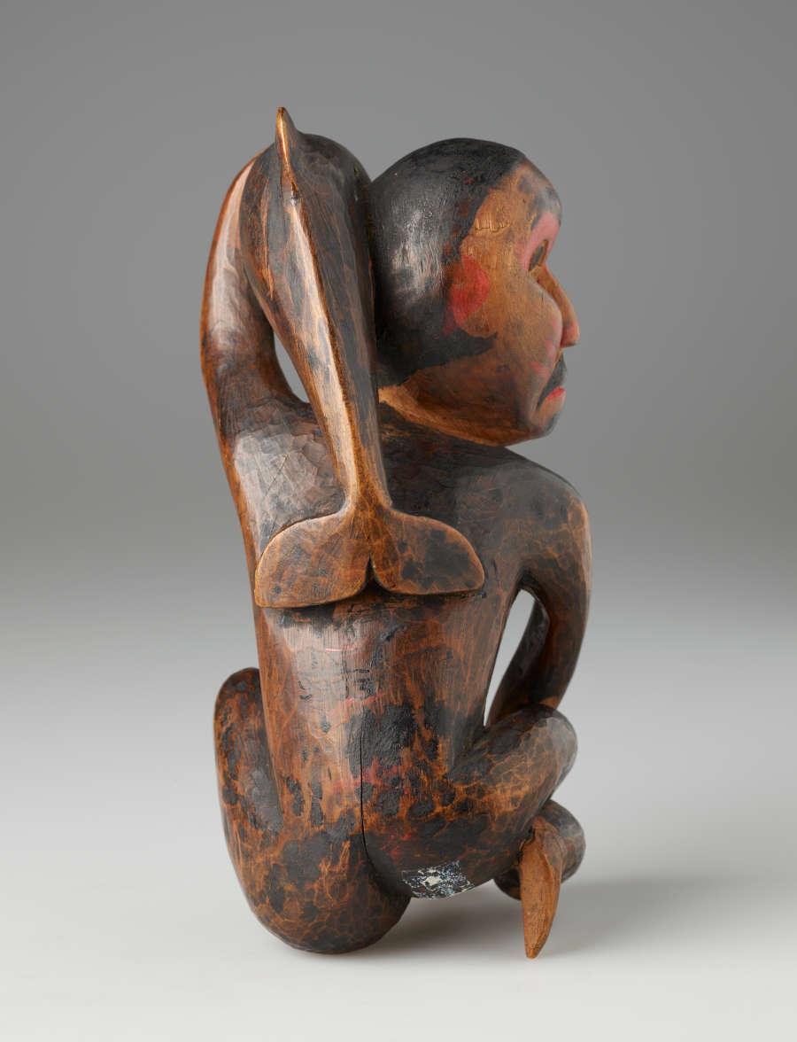 Back-view of worn painted wooden sculpture of a kneeling man looking aside, holding a small whale against his head. Visible is the whale’s tail resting on the man's back.