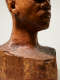 Dramatically lit, close-up view of the base and neck of a wooden-carved bust depicting a figure with afrocentric facial features. The material is textured while retaining gloss. “Elizabeth Prophet” is carved into the side.