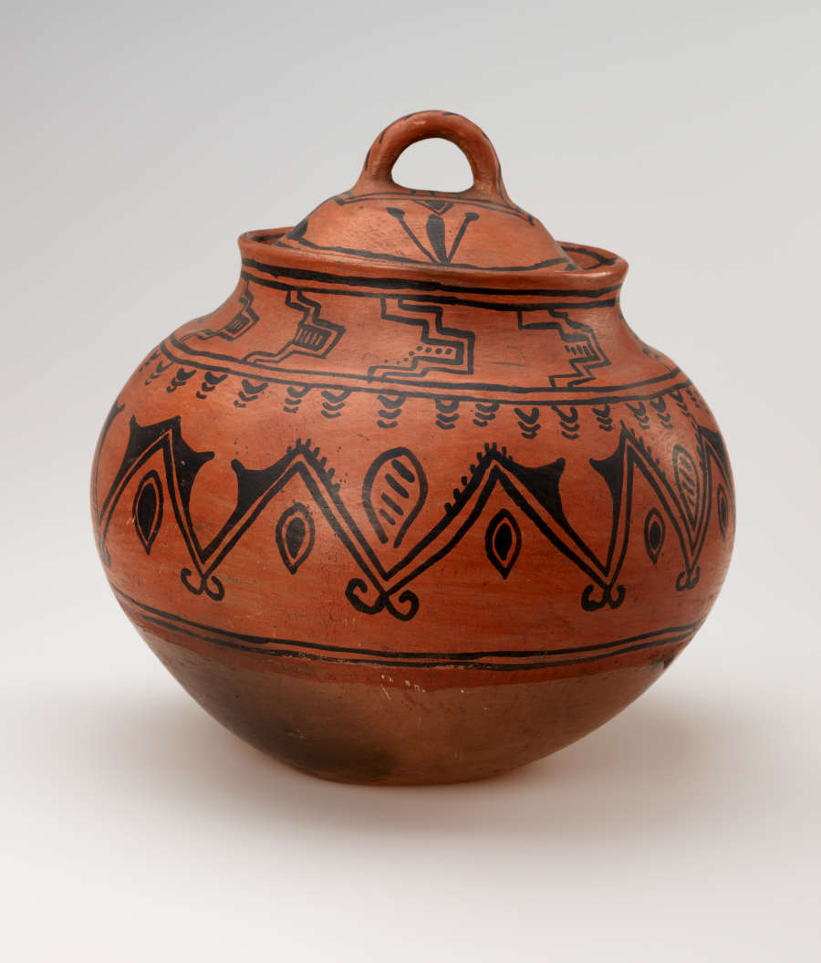 A bulbous red vessel with black abstract designs, leaning slightly to the right. The lid has a small round handle.

