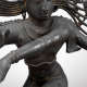 Detail of the figure showing one of the figure’s arms gesturing diagonally towards the viewer. The figure’s neck bears rows of necklaces, and the wrist and upper arms feature bands.
