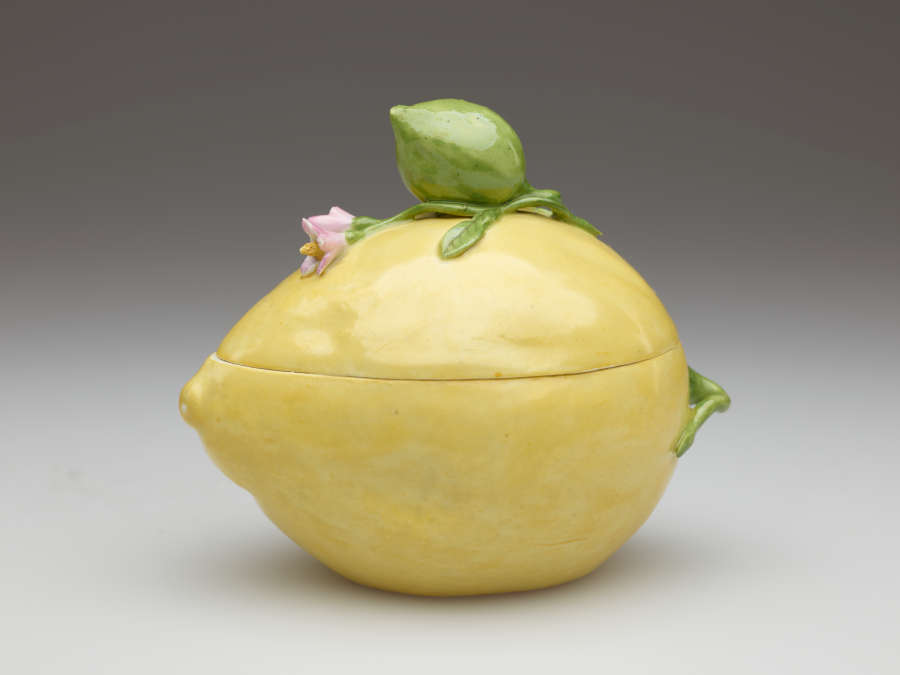 Lidded yellow box shaped like a lemon. Molded green leaves with a large bud and small pink flowers are applied to the top and wrap around the side.
