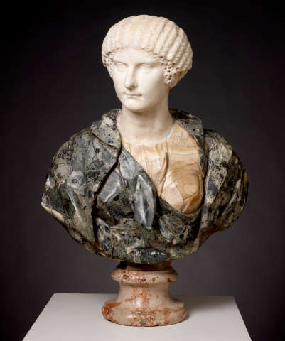 Realistic marble bust depicting a stoic woman whose clothes contrast with the smooth white marble of the bust, the outermost layer being textured black and gray stone, and the innermost tan and striped.