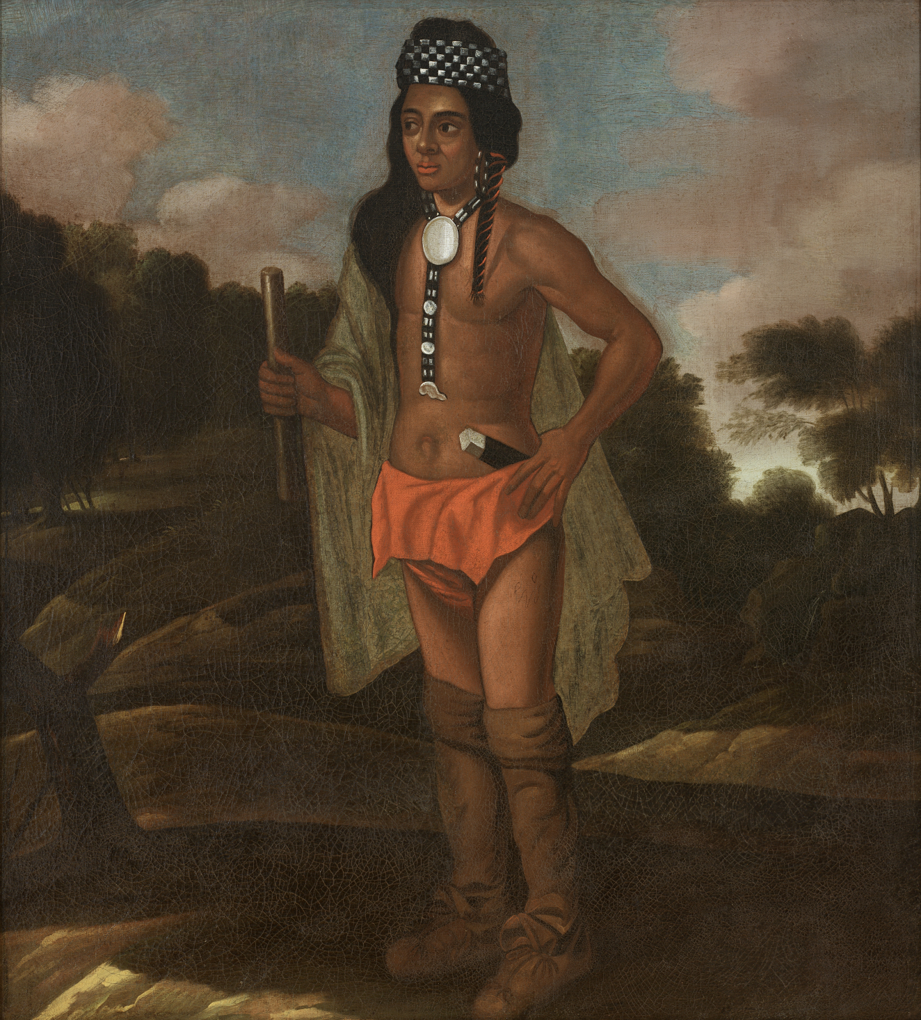 A brown skinned man with long hair, wearing a headdress, a beaded medallion, and carrying a short wooden staff stands in a dark landscape, looking into the distance.