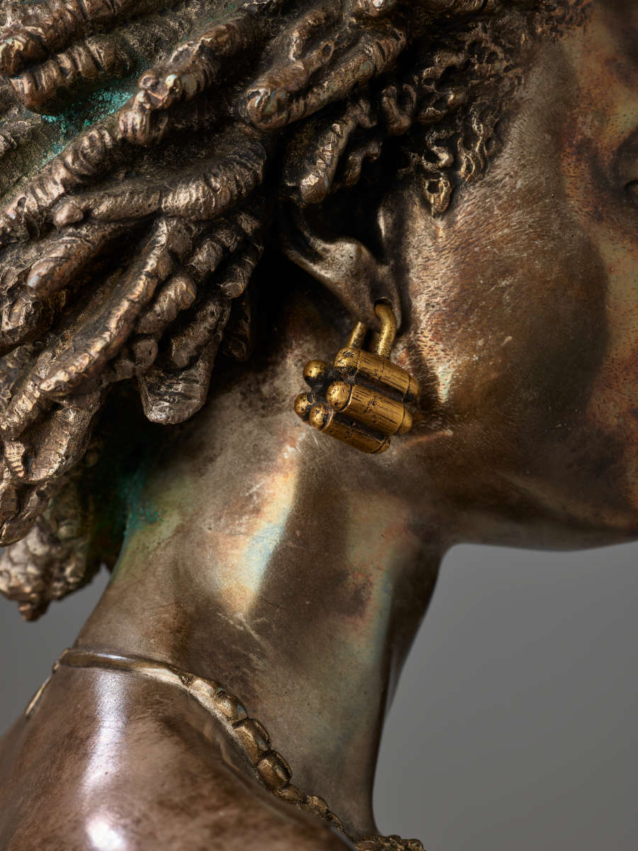 Close-up of right bronze cheek and ear displaying a chunky gold earring of clustered, bullet-shaped forms dangling near dark bronze intricate coiled hair.