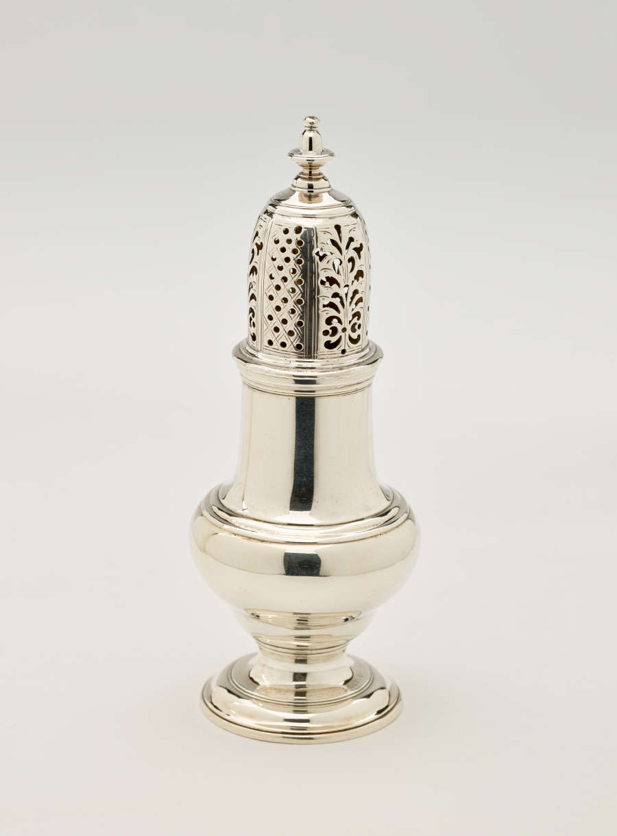 A silver vessel with a foot, rounded body that goes to a cylindrical top half, and the lid is perforated with small holes and floral cutouts.