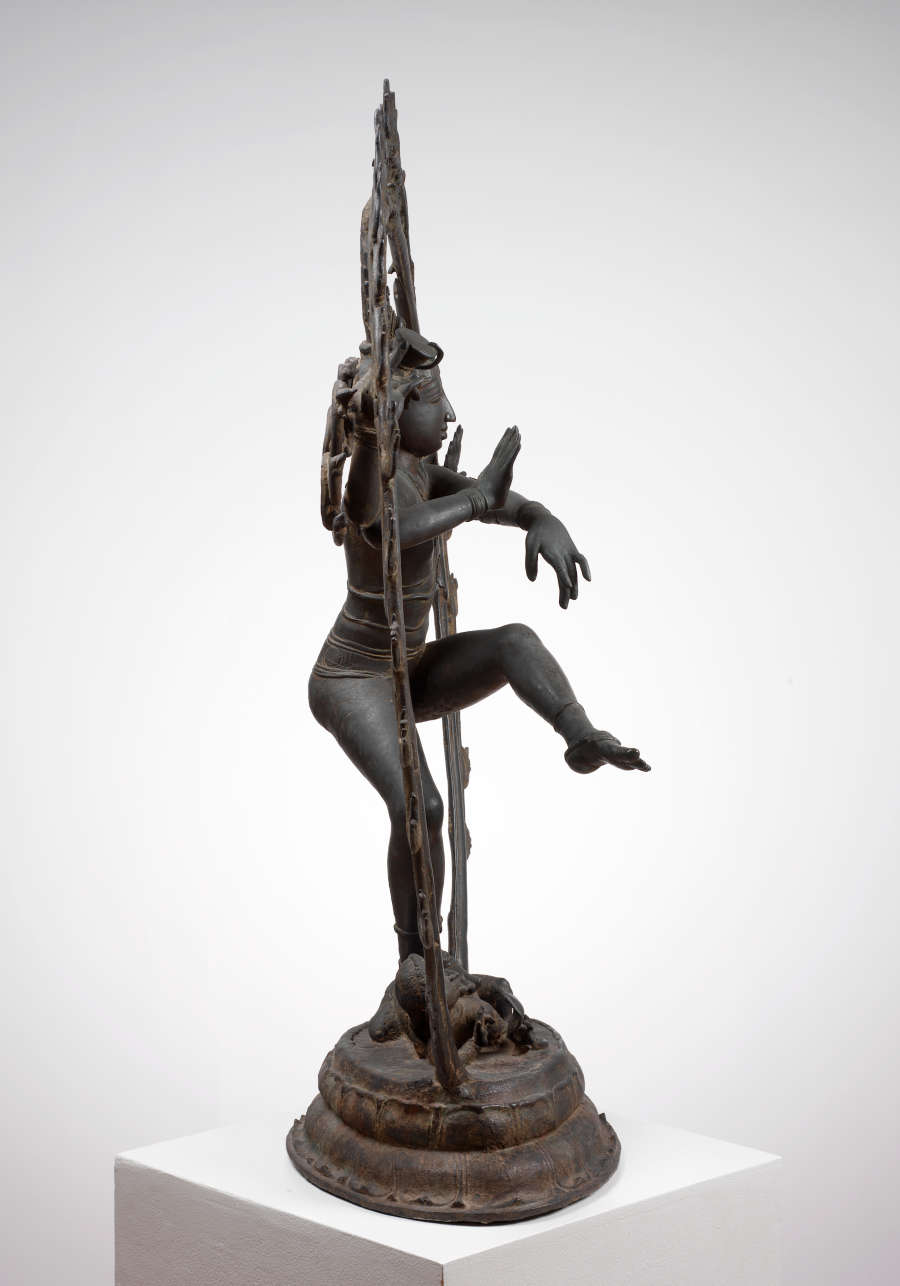 Side view of the bronze sculpture, showing the thin sculpted halo surrounding the figure and a truncated base. Two of the figure’s arms gesture outwards.