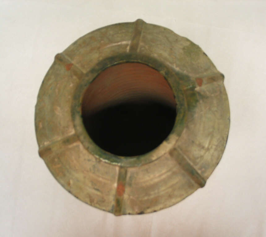 Top-view of a worn green-gray terracotta urn showing its narrow circular mouth flaring into its wide and shallow saucer-like neck. 6 ridges radially branch off from the mouth's edge.