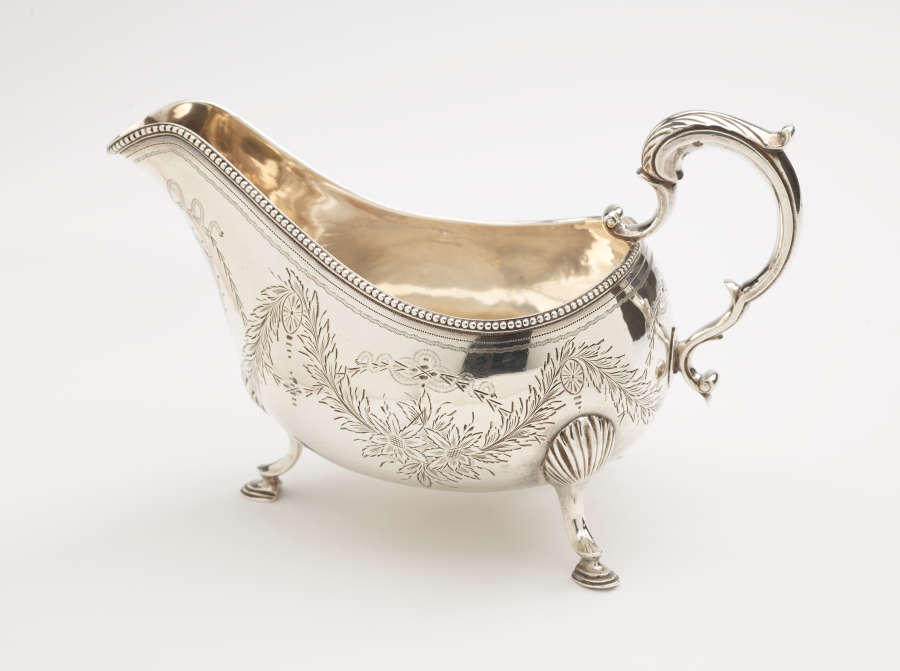 A silver sauce boat with a sculptural handle, high lip for pouring, and three protruding feet. There are floral engravings along the body of the vessel.