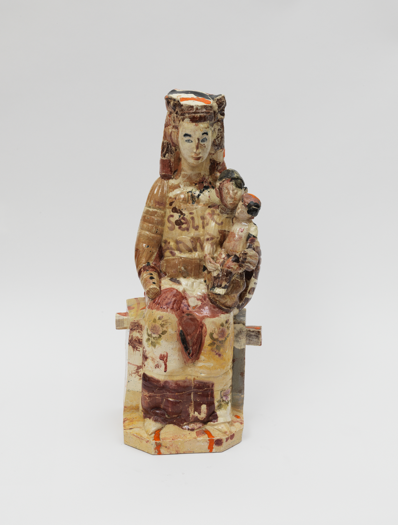 An intricately painted ceramic figure of a seated woman wearing a headdress cradling a small child in her left arm, the surface richly patterned with beige, red, creams, and browns.
