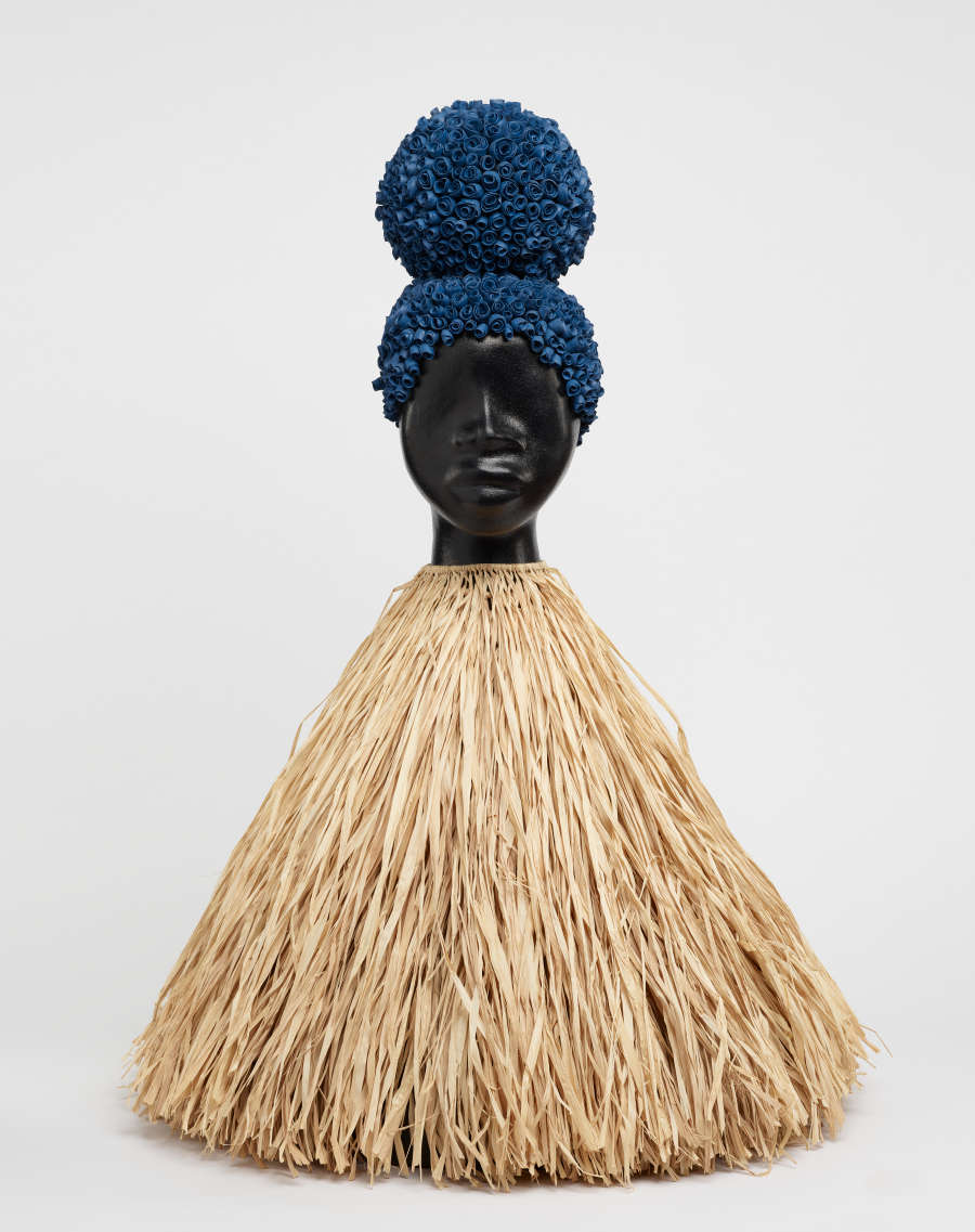 A sculpture composed of a dry husk tassel topped with a black glazed terracotta head. The head has hair in a bun made of folded woven dyed dark blue husks.