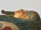 A gilded coffin lid with a gilded face, with blue-outlined eyes, a false beard, and a headdress, with gold detailing and visible signs of age and wear.