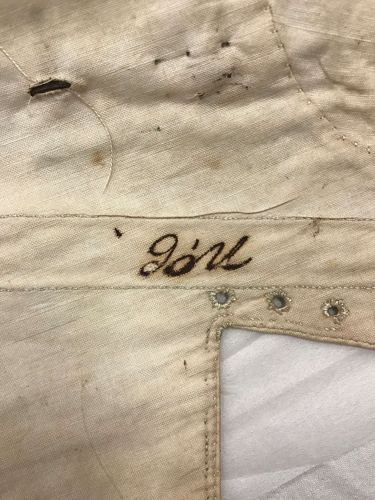 A close-up of an aged, cream-colored fabric with a handwritten word in dark ink on the side, worn embroidery, small dark stains, holes, and a small row of eyelets.