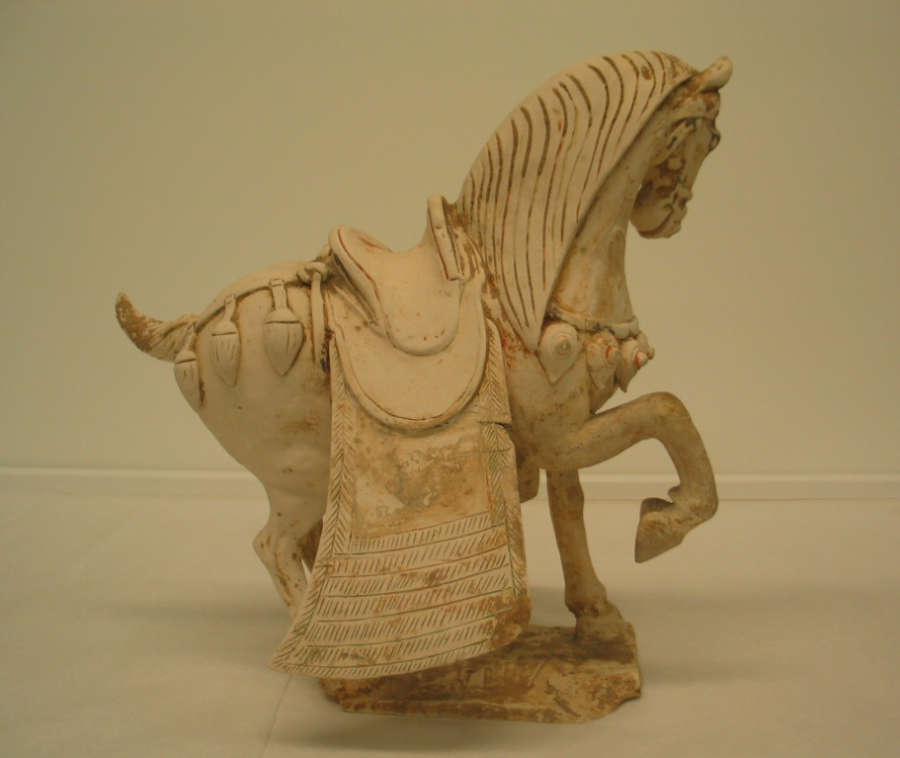 Another side-view of a white ceramic sculpture of a saddled, ornamented horse with one foot raised, a short wound tail and heavy mane. It stands on a thin connected base.