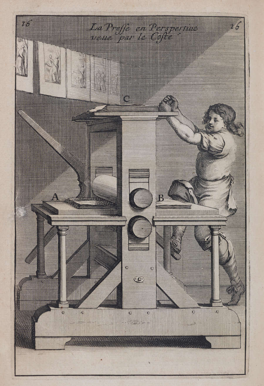 A printer operates a large wooden printing press while prints hang to dry above.