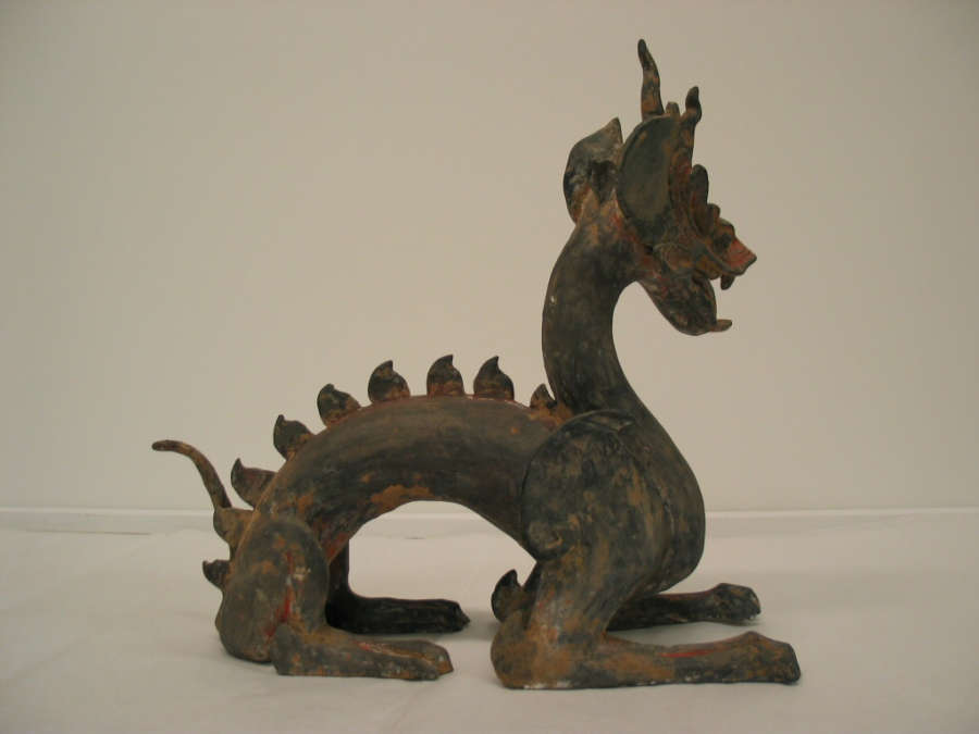 Side-view of an arched four-legged dragon with a lean body, open mouth, horned head, scaly back and a thin tail. The paint has worn into patches of browns and reds.