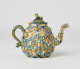 A teapot with a shell shaped body, decorative spout, and animal sculpted finial for the lid. It is mottled blue, brown, and cream in color.