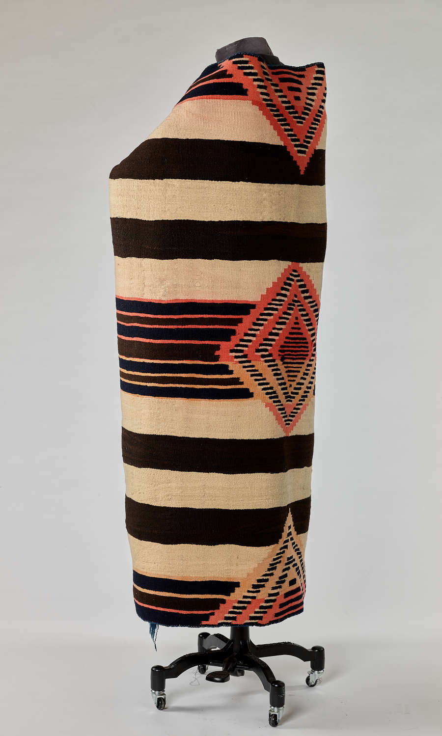 Back side view of a black and cream woven blanket with salmon pink and yellow geometric patterns draped around a mannequin. Three geometric shapes line down the center back symmetrically.
