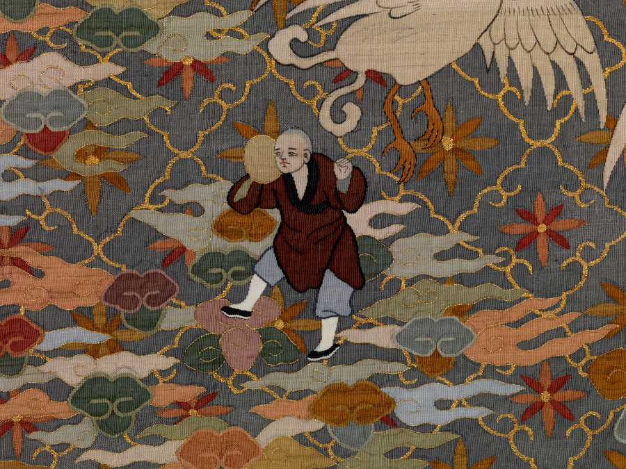 Robe detail showing a robed monk carrying an egg, amongst earthy pastel clouds. The background is dark with a golden diagonal grid, within each square is a floral motif.