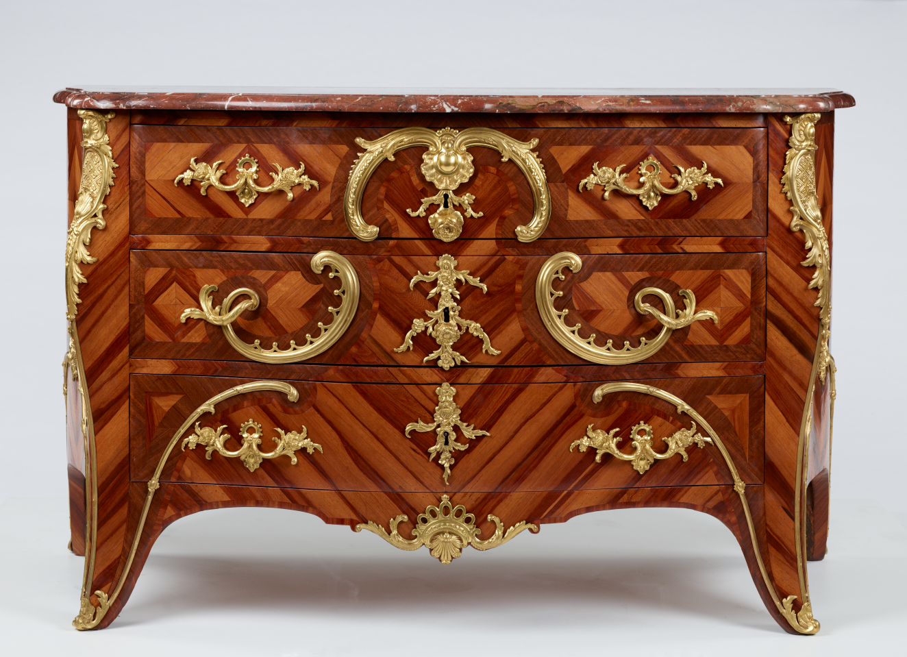 An antique reddish-brown wooden dresser with three drawers and intricate swirling gold detailing. The top surface is composed of red marble with white veining. 