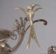 Detail of golden bird with long two-part tail and long curved beak perched on an ornate silver decorative scroll attached to a shallow dish above a partially visible figure’s face. 