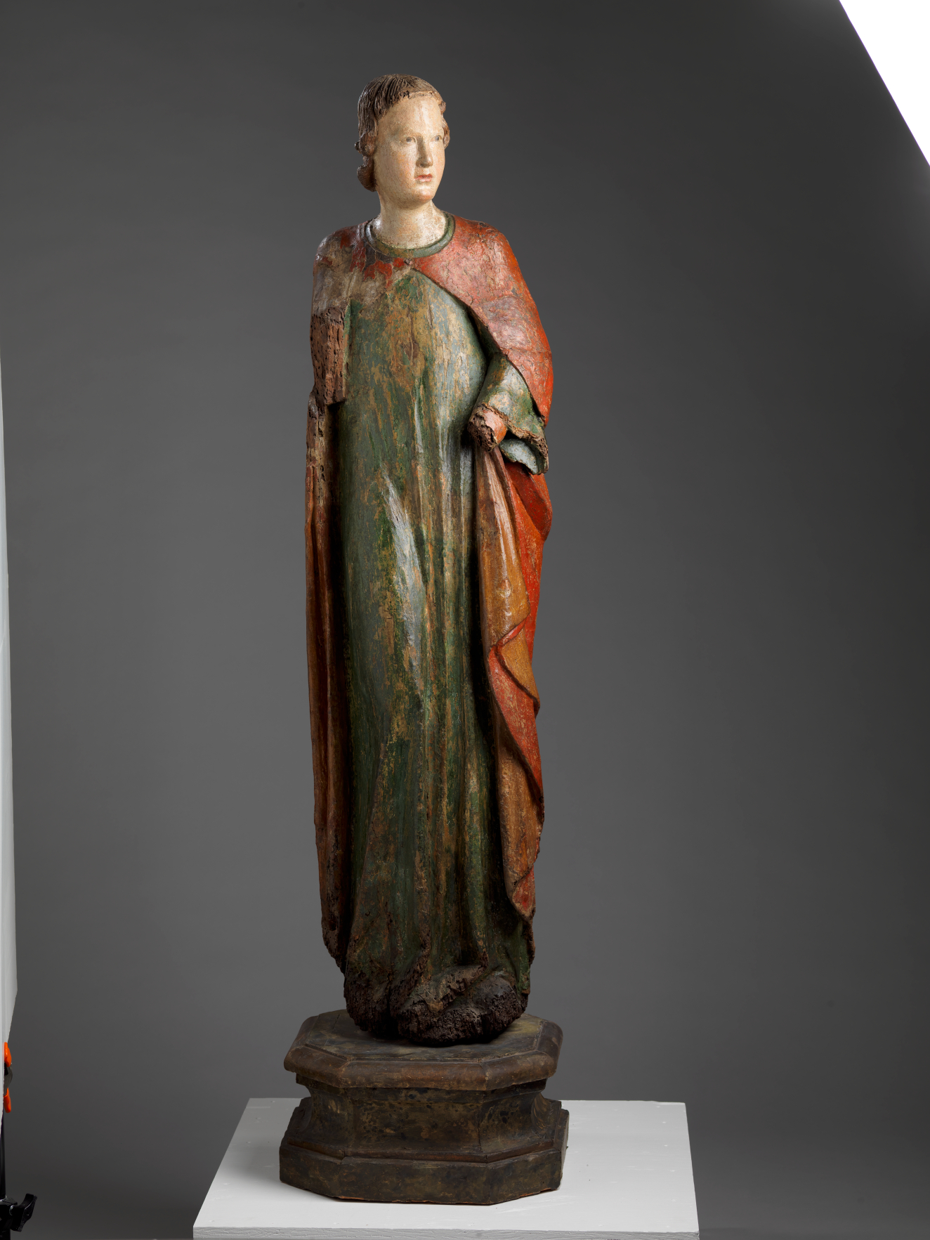 A tall, weathered wooden statue of a robed figure with a serene expression, draped in green and red garments, standing upon an octagonal base.