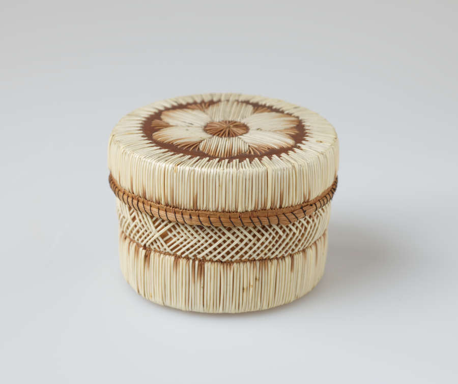 Cylindrical lidded leather box with cross-hatching and linear stripe patterns woven in white and brown straw. The brown rimmed lid has a central floral motif woven atop it.