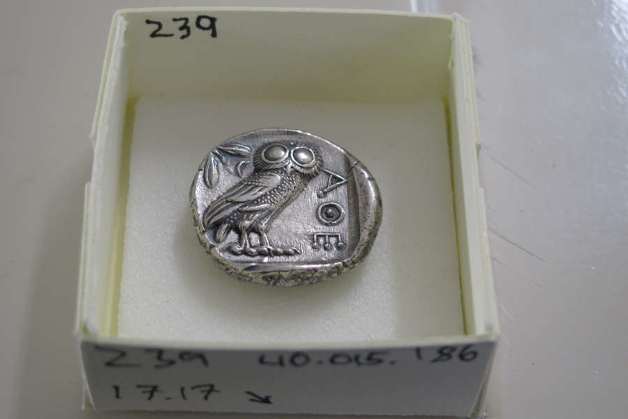Back of a round silver coin, stamped with an image of the Athenian owl, and the Greek letters ‘AOE’ beside it. It’s placed in a white box in storage labeled ‘239 40.015 186 17.7’.