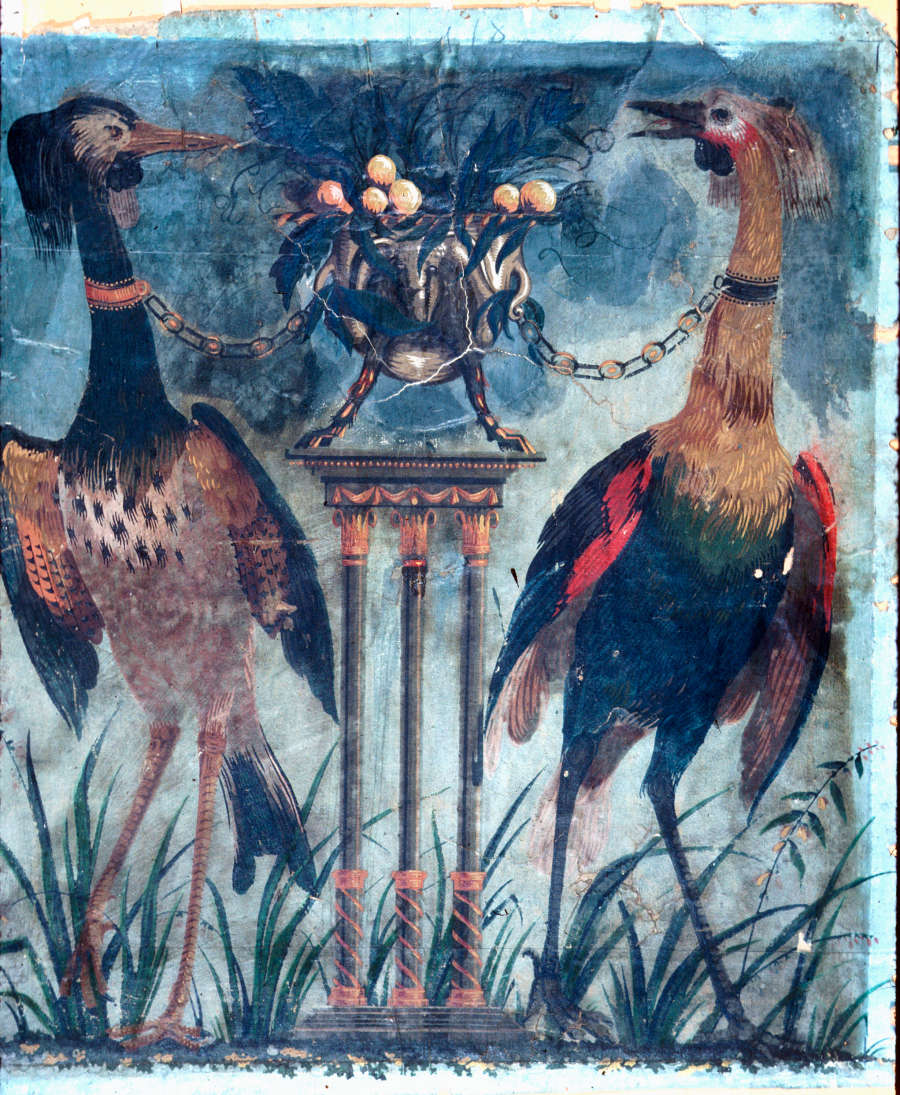 Ripped and weathered, cream painting of two large multicolored birds chained by the neck to a metal, fruit filled, vase on top of a three-legged pedestal.