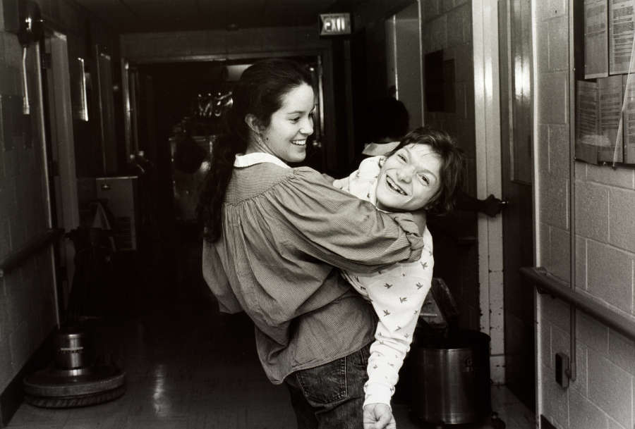 A grayscale photograph of a light-skinned woman standing in a hallway, a light-skinned girl in her arms. The girl looks towards the viewer laughing while the woman smiles at her.