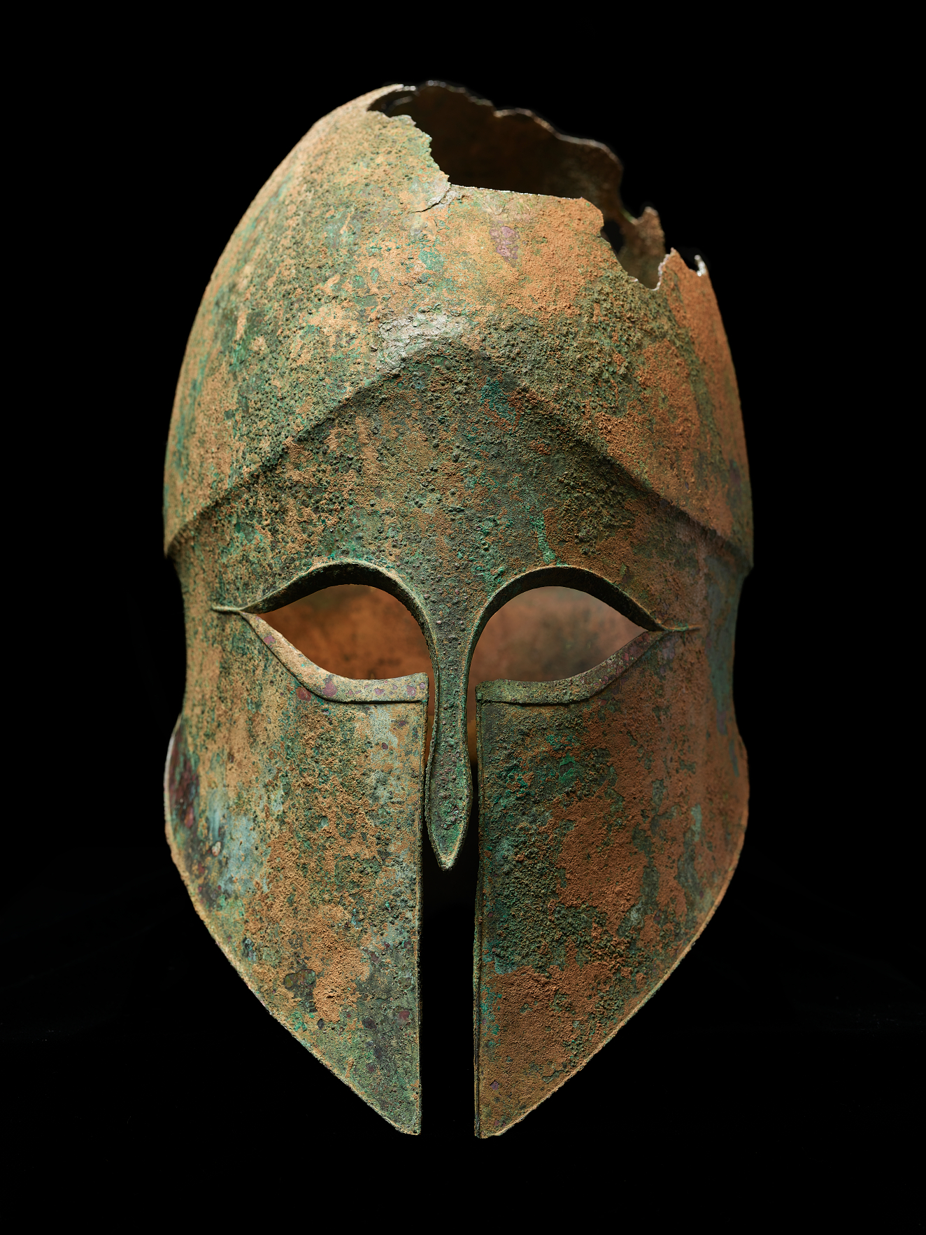Bronze helmet with a narrow nose guard and sharp eye holes. The corroded surface is brown with significant green discoloration. The top has jagged sections missing along the crown.