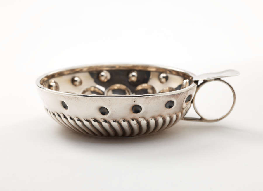 A silver, shallow vessel with a simple circular handle. Ribbed and circular decorations on the body.