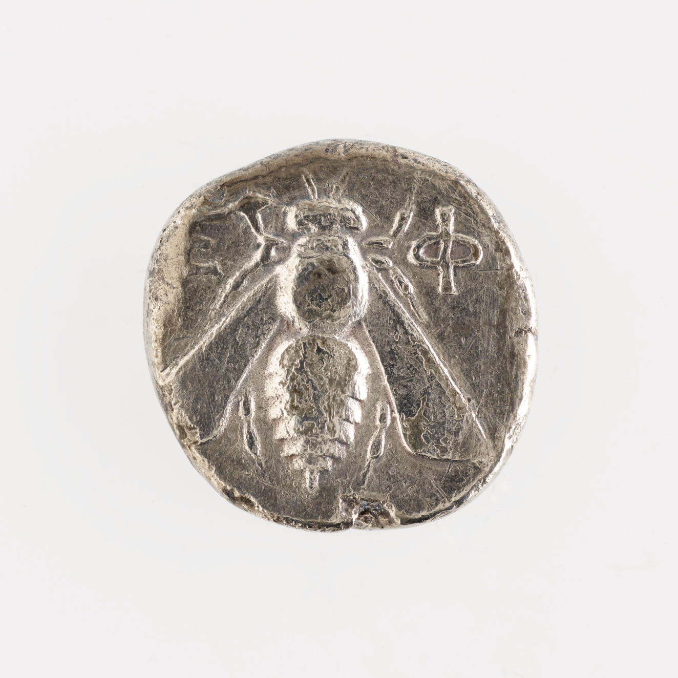 Rough-edged round silver coin embossed with an image of a bee with Greek letters beside it.