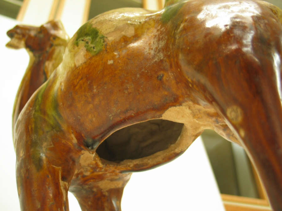 Detail view of the underbelly of a camel figurine, showing the glossy and worn green, tan, and brown surface and the hole in its stomach.