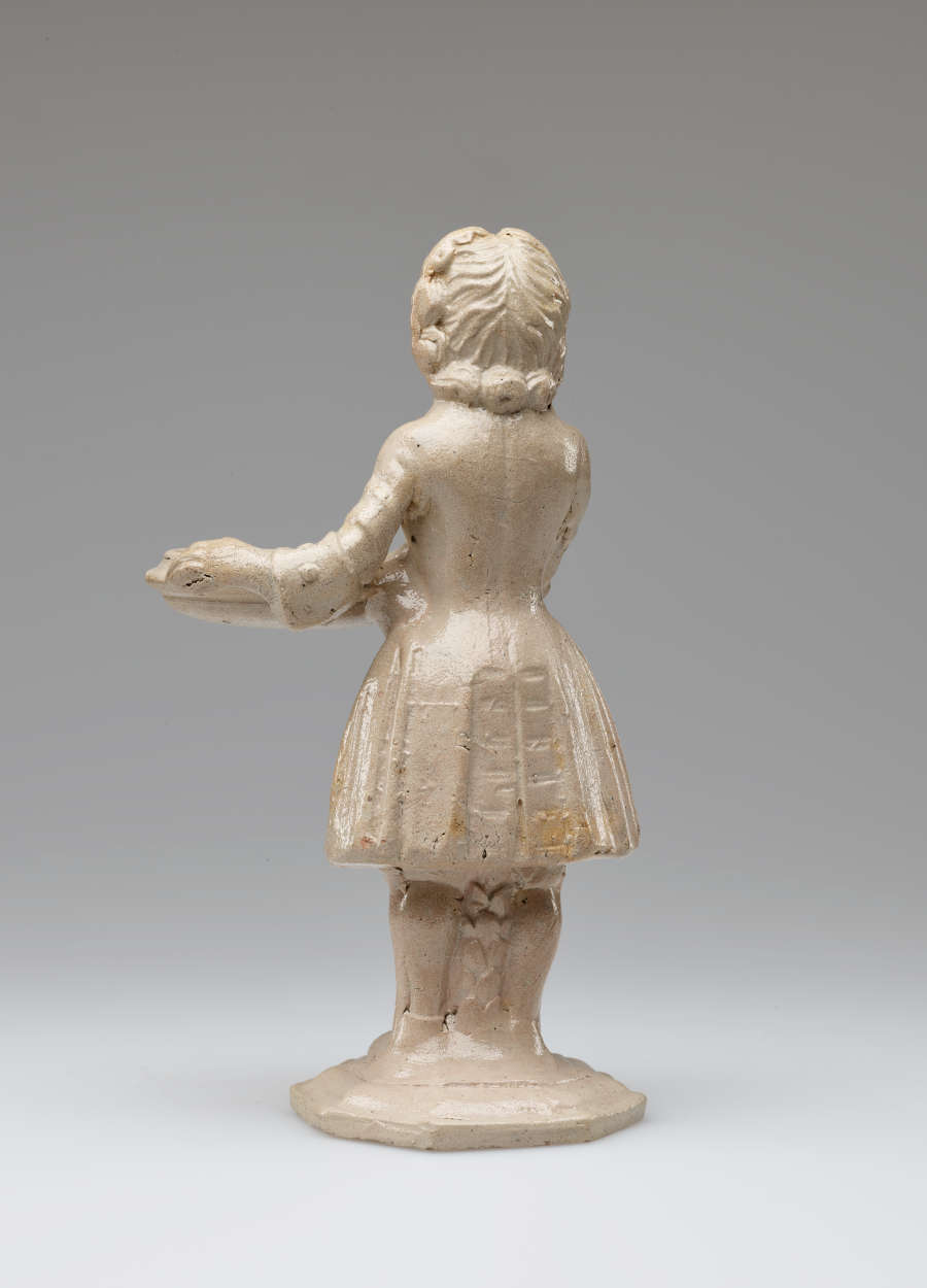 A cream colored sculptural figure holding a vessel, dressed in historical clothing with tightly curled hair.
