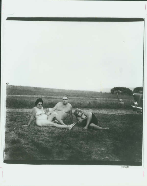 Black and white photo of an awkward, naked, light-skinned family reclined on grass. The lumpy woman only wears sandals. The equally lumpy man appears naked. The adolescent child wears shorts.
