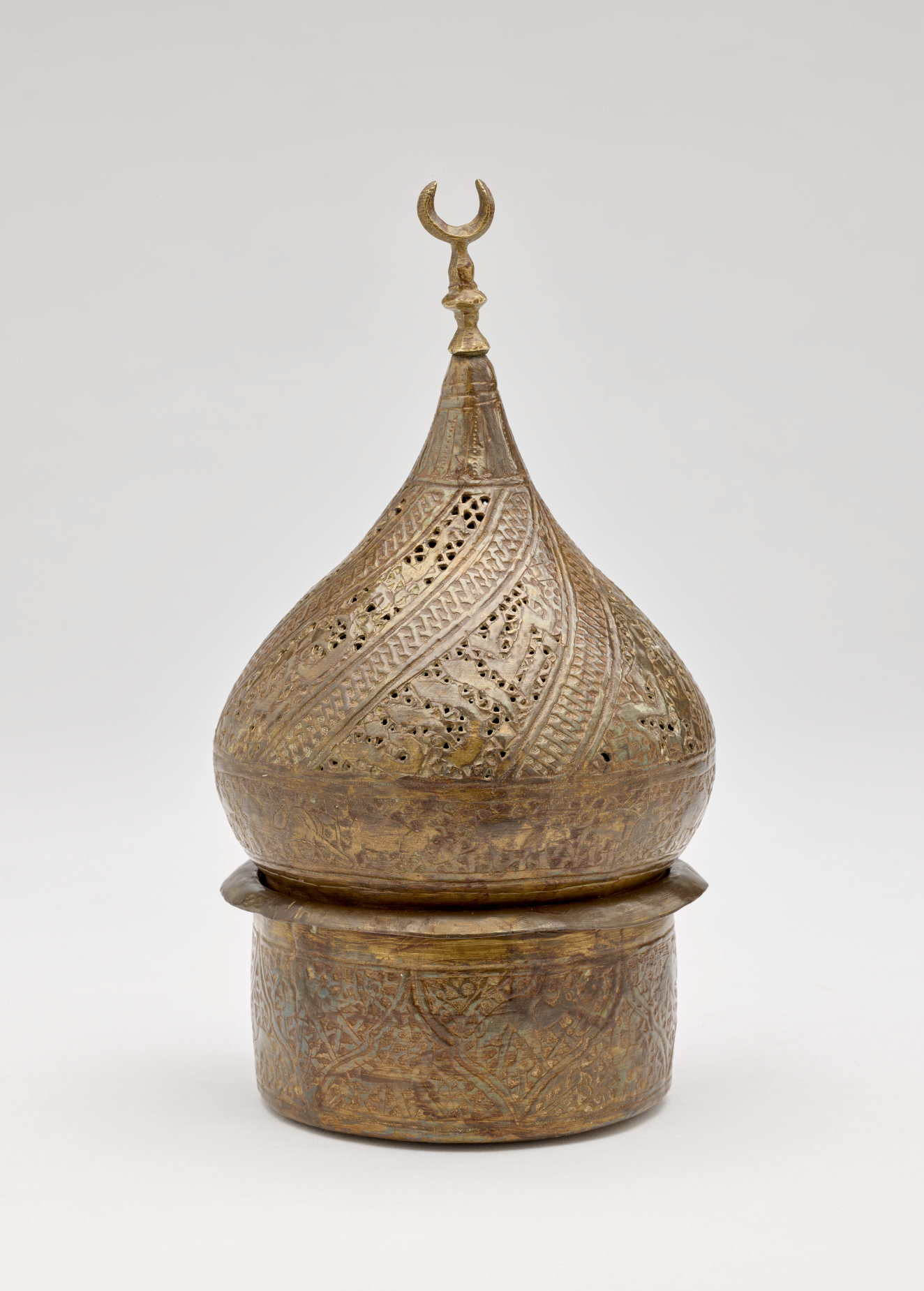 Bronze-colored sculptural object with incised design. Top half is teardrop shaped with a crescent moon shape on top. Cylindrical vessel on bottom.