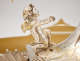 Detail of silver cherub in a leafy crown with small gold wings gazing upwards with outstretched arms, kneeling with one knee up at the base of a curved silver surface. 